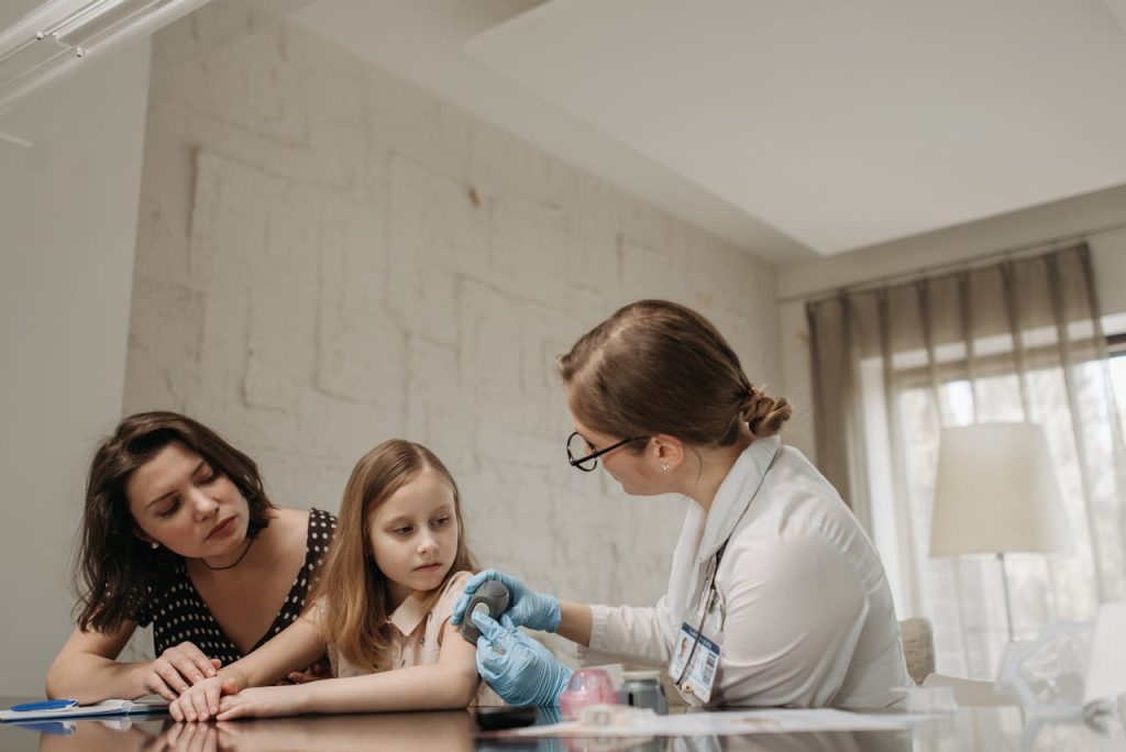 Mãe jovem branca está com sua filha, criança, branca, sendo atendidad por uma médica, branca, jovem. A médica mostra um instrumento para a criança e explica a ela e sua mãe.